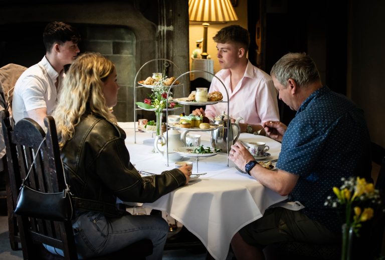 Four people are seated around a table having afternoon tea at Holdsworth House. The table is set with a white tablecloth, teacups, saucers, a teapot, and a tiered stand filled with various pastries and desserts. On the left, a young woman with blonde hair, wearing a leather jacket, sits next to a young man in a white shirt. Across from them, an older man in a blue shirt and a younger man in a pink shirt are seated. The setting is cozy with dim lighting, a lamp in the background, and floral decorations on the table.
