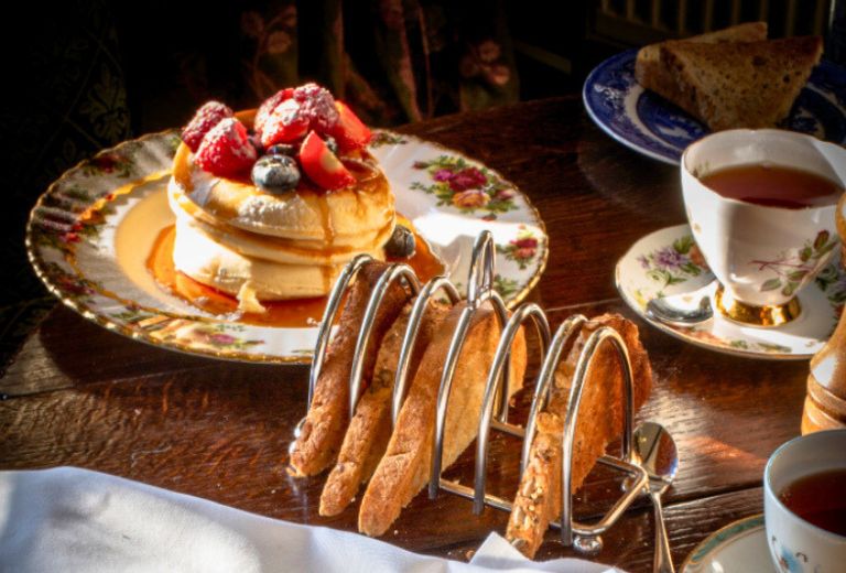 A plate of fluffy pancakes topped with fresh berries and syrup, served with tea in fine china and a toast rack on a wooden table.