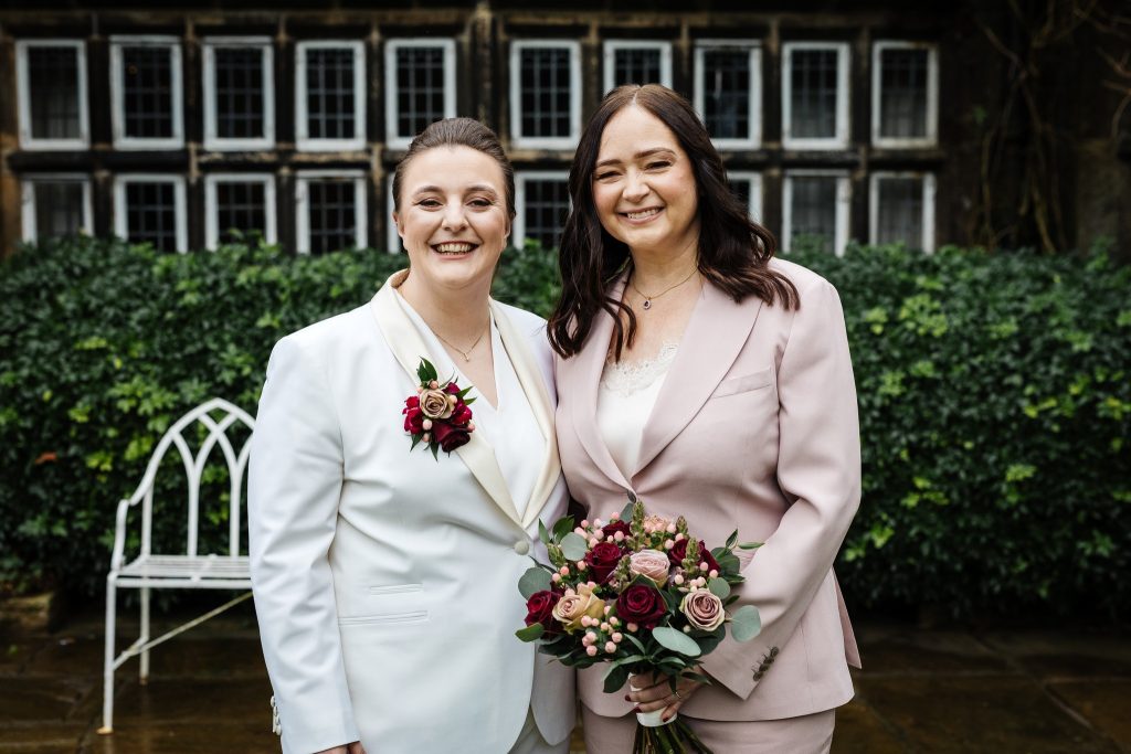 Smiling same-sex couple in tailored suits holding a bouquet in front of the historic façade of Holdsworth House