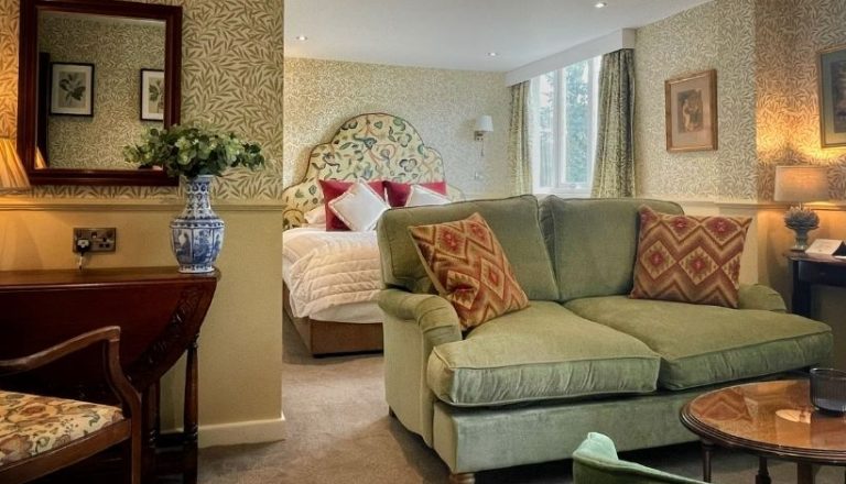 A cozy hotel suite with a green sofa, floral wallpaper, and a comfortable bed in the background, elegantly decorated with cushions and artwork.
