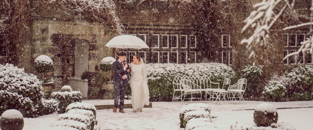 Bride and groom walking under an umbrella through snow-covered gardens outside a historic manor house.