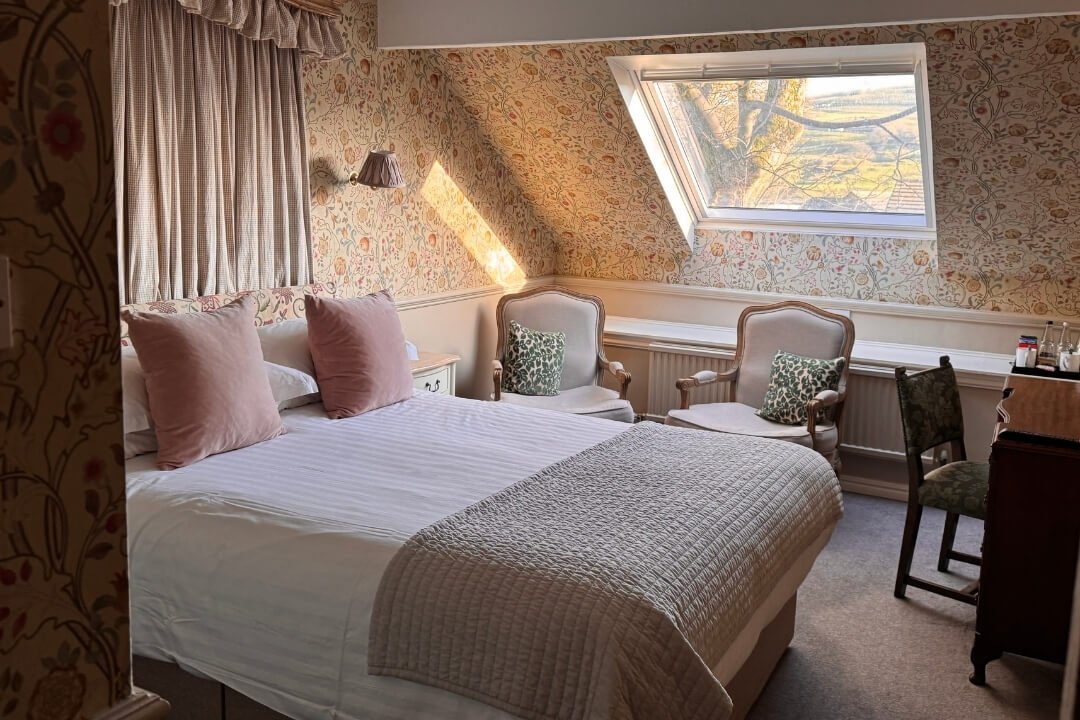Cosy hotel bedroom with floral wallpaper, a large bed, armchairs, and a slanted window overlooking the grounds.