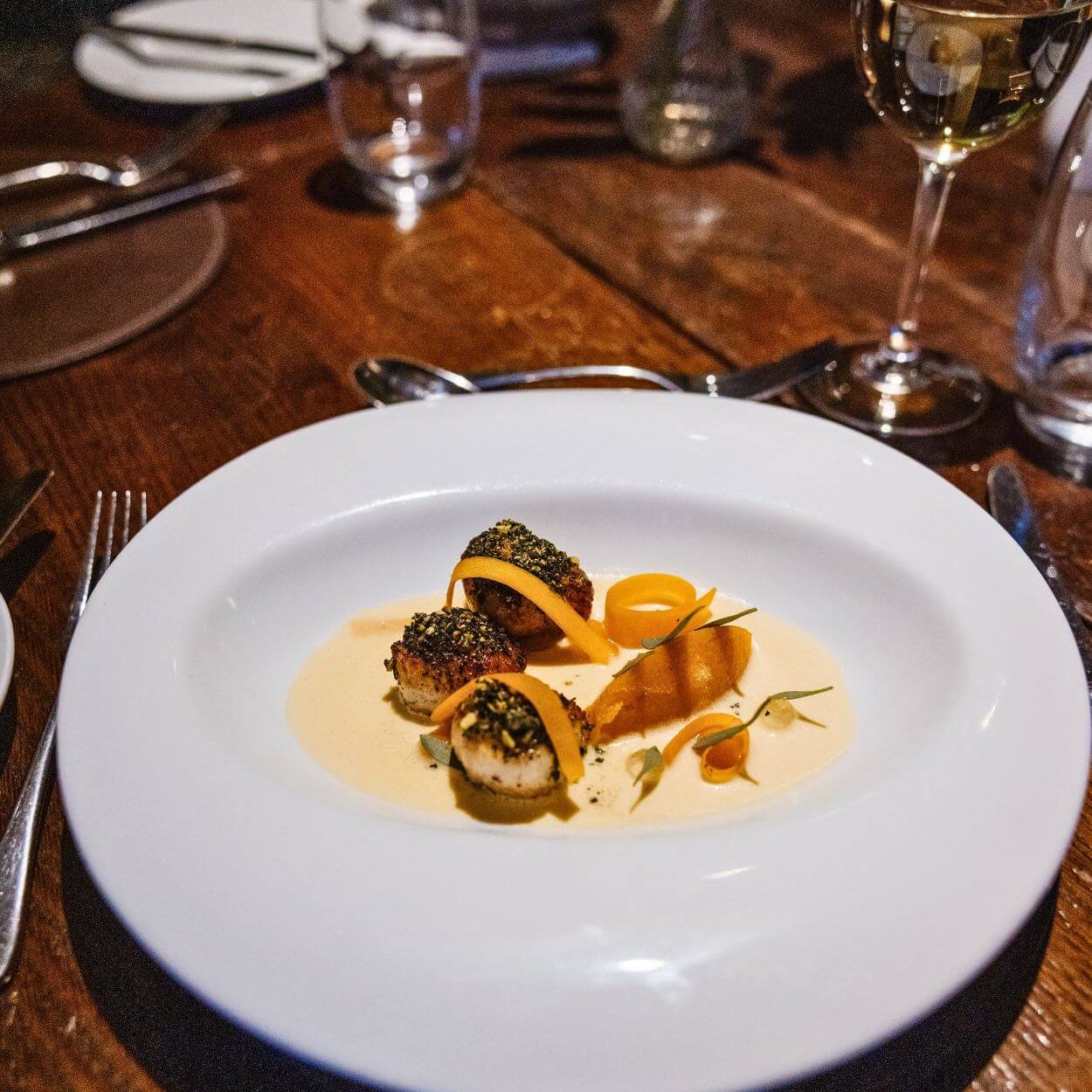 Plated à la carte dinner at Holdsworth House featuring seared scallops with seasonal garnishes, served on a white plate on a wooden restaurant table.