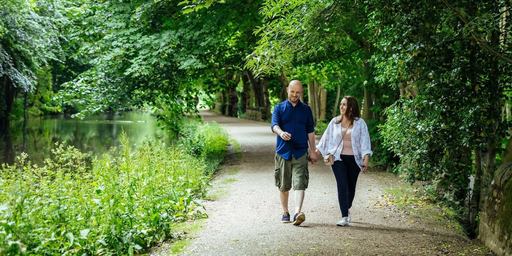 A man and woman holding hands walking alongside a river surrounded by lush green forest in Saltaire, Bradford.